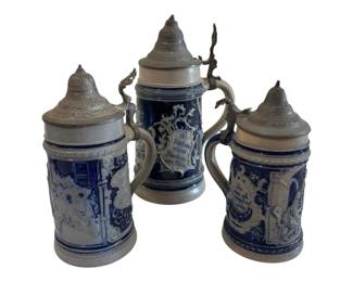 556 - 3 German Blue Salt Glaze Steins 9.5" (Description): Collection of three German-style salt glaze  stoneware steins with pewter lids. Each stein  features traditional cobalt blue decoration  with relief scenes depicting figures in  tavern settings, surrounded by ornate scrollwork and German text cartouches. The  steins have curved handles and decorative  beehive-shaped pewter lids with thumblifts.  One stein is broken. Measures 9.5 inches  tall.