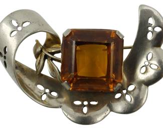 1914 - Sterling Art Deco Brooch 1.21oz (Description): Sterling silver Art Deco brooch featuring an  elegant design with pierced floral motifs and  a large faceted amber-colored stone. The  brooch showcases classic Art Deco styling  with flowing ribbon-like curves and delicate leaf accents. Weighs 1.21 ounces.