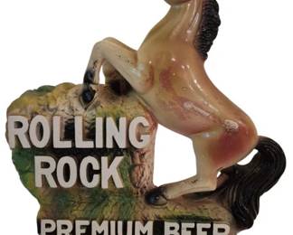 3940 - Rolling Rock Beer Horse Advertisement (Description): Vintage Rolling Rock Premium Beer  advertisement featuring a rearing horse  figure atop a naturalistic rock base. This  chalkware or plaster bar display piece  showcases the iconic Rolling Rock branding with raised white lettering reading "Rolling  Rock" and "Premium Beer" against a painted  green and brown rocky backdrop. The horse is  painted in tan and cream tones with a black  mane and tail. Measures 12" x 11".