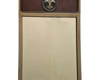 1316 - Decorative Mirror w/ Fleur-de-Lis 15"x12" (Description): Decorative wall mirror featuring an ornate  fleur-de-lis medallion accent. The frame has  a distinctive curved design on one side with  a brown leather or faux leather panel  bordered by rope-style trim. The mirror portion is set within a gilt-toned frame.  Measures 20" x 12".
