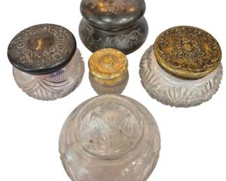 2715 - Assortment Vintage Dresser Boxes (Description): Some featuring cut glass bodies with  decorative metal lids. The assortment  includes various sizes and styles, with lids  in silver-toned and gilt finishes displaying  ornate repousse floral and scrollwork designs. One jar features an engraved  monogram on its domed silver lid. The glass  bases showcase different cut patterns  including hobnail, swirl, and starburst  designs, typical of Victorian-era vanity