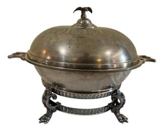585 - Vtg Silver Plate Covered Serving Dish w Eagle (Description): Vintage silver plate covered serving dish  featuring an eagle finial on the domed lid  (wing broken). The lid is decorated with  elegant engraved foliate and medallion  designs. The piece includes a footed warming stand with ornate scrolled legs ending in paw  feet, featuring a reeded band detail. Wing  damaged. Four pieces total. Measures 9" x  13".