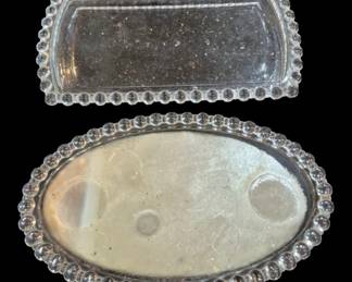 1975 - Two Glass Vanity Trays w/ Beaded Edges (Description): Two glass vanity trays featuring decorative  beaded edges. The set includes one  rectangular tray and one oval tray, both with  mirrored or silvered glass surfaces  surrounded by raised glass bubble or bead borders. Suitable for displaying perfume  bottles, jewelry, or other vanity  accessories.