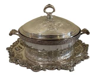 590 - Silver Plated Covered Serving Dish w/ Tray (Description): Silver plated covered serving dish with  matching tray. The domed lid features  embossed floral decoration and a ring finial  handle. The round bowl displays ornate  engraved scrollwork and floral patterns with twin scrolled handles. Sits on an elaborate  footed tray with shell and scroll border  design in the Rococo style. Measures 8" x  12".