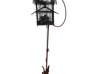 3739 - Decorative Bird Cage on Wrought Iron Stand (Description): Decorative bird cage on stand featuring an  ornate pagoda-style cage with tiered roofline  and vertical bars. The cage is mounted on a  tall wrought iron stand with a curved hook  arm and scrolled tripod base with decorative leaf motifs. The cage is adorned with  feathers at the top.