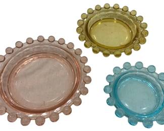 988 - Tri Colored Glass Candlewick Coasters Set (Description): Set of three colored glass coasters or  cellars featuring a distinctive candlewick  beaded edge design. The collection includes  one pink, one amber/yellow, and one blue  coaster, each with a smooth center well surrounded by a decorative ball-edge border.