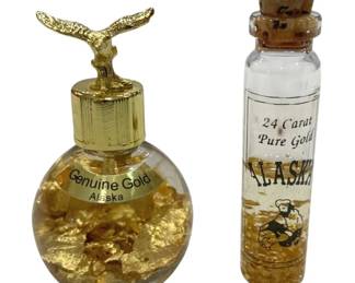 1204 - 2 Alaska Gold Flake Souvenir Bottles 2.5" (Description): Two decorative Alaska souvenir bottles  containing gold flakes. The first is a round  glass bottle labeled "Genuine Gold Alaska"  with a gold-tone cap topped by a whale tail  finial. The second is a cylindrical cork-topped vial labeled "24 Carat Pure Gold  Alaska" featuring a gold prospector  illustration. Both bottles are filled with  gold flakes suspended in liquid.