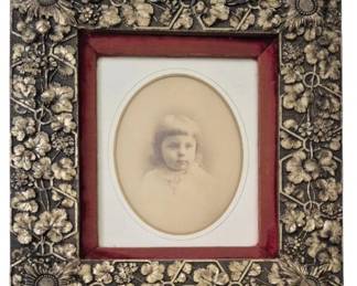 1276 - Vtg Floral Repousse Picture Frame (Description): Vintage decorative picture frame featuring  elaborate floral repousse work with  sunflowers, leaves, and ribbon motifs  throughout the border. The frame has a red  velvet inner liner surrounding an oval opening. Contains an antique sepia-toned  portrait photograph. Measures 17"x15".
