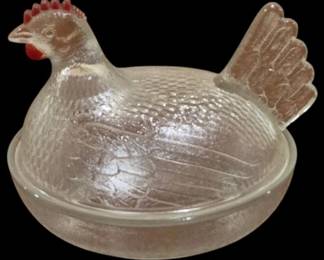 4037 - Crystal Hen on Nest Covered Dish Red Trim (Description): Crystal hen on nest covered dish with red  painted accents on the comb, wattle, and tail  feathers. The pressed glass piece features  detailed feather texturing throughout the  body and a nest base with visible eggs beneath the hen. A charming country kitchen  collectible. Measures 5 1/2" x 7".