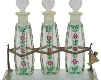 859 - Vintage Painted Perfume Set on Stand 7"x7"x2" (Description): Vintage perfume set featuring three  decorative glass bottles with faceted crystal  stoppers, displayed in a metal holder on a  mirrored base. Measures 7 inches by 7 inches  by 2 inches.