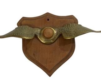 2762 - Antique Brass Winged Radiator Cap on Wood (Description): Antique brass winged radiator cap mounted on  a wooden shield-shaped plaque. The decorative  brass piece features detailed wing designs  extending outward from a central circular  element, displayed as a wall-mounted trophy or decorative accent.