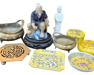 1422 - Asian Decorative Collection w/ Figurines (Description): A collection of Asian decorative items  including a glazed ceramic mudman figurine of  a seated elder on carved wood stand, a white  porcelain Guanyin figure, two brass footed  censers or bowls, a brass trivet with geometric shou symbol design, a yellow ground  famille jaune porcelain covered box with  floral decoration, a matching yellow floral  porcelain dish, and a yellow ground porcelain  plate with pink peony motif and Greek key