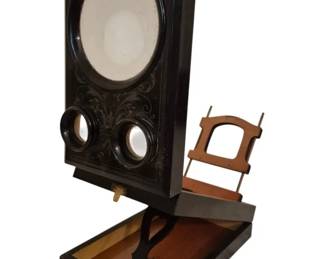 4011 - Vintage Stereo Graphoscope Viewer (Description): Vintage stereo graphoscope featuring an  ebonized wood frame with decorative foliate  engravings on the front panel. The viewer is  equipped with a large circular magnifying  lens above two smaller stereoscopic viewing lenses. The unit is mounted on a hinged  storage base with wooden interior  compartments, designed to hold stereo cards.  A wooden card holder arm extends from the  base for positioning viewing materials at the