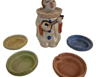 4185 - Oriental Made in Japan Smoking Set (Description): An Oriental smoking set made in Japan. The  set features a figural clown playing a  stringed instrument that serves as the  centerpiece, accompanied by four coordinating  ashtrays in various colors including yellow, green, blue, and brown.