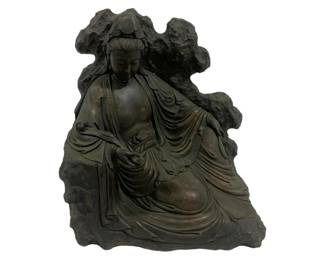 8003 - Bronze Guanyin Bodhisattva Statue (Description): Bronze statue depicting Guanyin, the  Bodhisattva of Compassion, seated in a  relaxed royal ease pose against a  naturalistic rock formation backdrop. The  figure is draped in flowing robes with finely detailed folds and wears a traditional  topknot hairstyle. The bronze displays a dark  patina consistent with age. Measures 13.5  inches wide by 9.5 inches deep by 16 inches  tall.