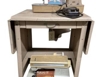 4086 - Metal Stand w/ 2 Singer Sewing Machines (Description): Metal sewing machine stand with two Singer  sewing machines. The lot includes a vintage  Singer sewing machine in beige/tan coloring  resting on the stand, along with a Singer  Futura II model with its original manual and foot pedal under the stand. The metal stand  features drop leaves on each side and rolls  on casters for mobility.  A box of sewing  accessories is also included. Used as is electric/electronic/mechanical