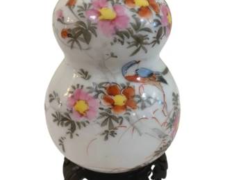 332 - Oriental Porcelain Double Gourd Vase w/ Stand (Description): Oriental porcelain vase in a traditional  double gourd form, featuring hand-painted  floral decoration with pink, yellow, and  orange blossoms among green foliage, accented  by a colorful bird perched on a branch. The white ground vase has gilt trim at the neck  and rests on a carved wooden display stand.  Measures 8.5" tall.