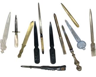 1201 - Group of Assorted Vintage Letter Openers (Description): A diverse collection of letter openers in  various materials and styles. The group  includes examples in crystal or glass with  carved fish motif, mother of pearl with  dagger-style blade, ornate brass with decorative handle, black bakelite or plastic  with figural designs, polished steel,  gold-tone metal, and a silver-toned example  with magnifying glass feature. The collection  represents a range of periods and decorative