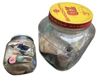4107 - 2 Vintage Glass Jars Filled with Buttons (Description): Two vintage glass jars filled with assorted  buttons. The larger jar features a yellow and  red OZ Smooth peanut butter lid, while the  smaller jar has a white lid. The collection  includes buttons of various sizes, colors, and materials, offering a diverse assortment  suitable for crafting or collecting.