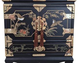 1278 - Black Lacquer Chinoiserie Jewelry Cabinet (Description): Black lacquered two-door cabinet in  Chinoiserie style featuring hand-painted  floral and bird motifs with gold accents. The  cabinet is adorned with decorative brass  hardware including corner brackets, hinges, and ornate door pulls with brown tassels. A  lower drawer provides additional storage. The  piece rests on bracket feet with a scalloped  apron. Measures 15" x 16" x 9".