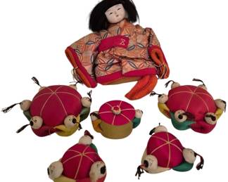 4184 - Vtg Japanese Fabric Pin Cushions w/ Doll (Description): Collection of vintage Japanese-style fabric  pin cushions featuring a larger seated doll  figure in an orange and red patterned kimono  with black hair, accompanied by six smaller  decorative pin cushions. Each small cushion features multiple tiny doll heads with black  hair arranged around pink silk cushions with  gold cord detailing. Also includes a small  round lidded container with matching pink and  gold design.