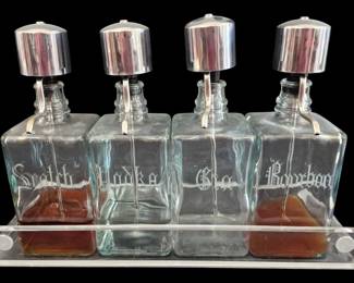 512 - 4 Glass Liquor Decanters w/ Acrylic Tray (Description): Set of four glass liquor decanters with an  acrylic holder tray. Each square decanter  features etched Old English script labeling  for Scotch, Vodka, Gin, and Bourbon. The  decanters are fitted with chrome pump-style pourers. Mid-century modern barware set in  good condition. Measures 11" x 17".