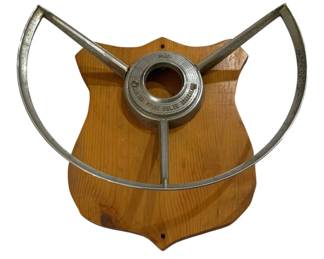 2761 - Ford Vtg Steering Wheel Mounted on Wood (Description): A vintage Ford steering wheel mounted on a  wooden plaque. The chrome steering wheel  features the classic three-spoke design with  a Ford center hub, displayed as a decorative  piece on a shield-shaped wooden backing.