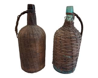 555 - Pr Vtg Wicker Covered Wine Bottles 15.5" (Description): A pair of vintage wicker covered wine bottles  with braided handles. One bottle features  dark amber glass with a brown cap, while the  other has aqua-tinted glass with a green  stopper. Both are fully wrapped in woven wicker casing with some age-related wear  visible. Measures 15.5 inches tall.