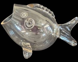527 - Blenko Clear Glass Fish Vase 7"x12.5" (Description): Blenko clear glass fish vase in the iconic  mid-century modern design. This charming  blown glass piece features the characteristic  rounded body with applied spiral eyes,  ribbed dorsal fin, sculpted tail fin, and small bottom fin. The open mouth serves as  the vase opening. The piece exhibits the  quality craftsmanship Blenko is known for  with smooth, clear glass and whimsical  artistic styling. Measures 7 inches by 12.5