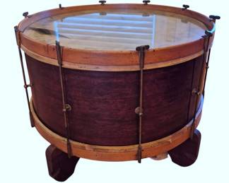 613 - Bass Drum Converted to Glass Top Table (Description): Antique bass drum creatively repurposed as a  glass top display table. The drum features a  rich mahogany-stained wooden shell with  natural wood hoops at top and bottom, secured  by traditional hook-style tension rods with T-handle tuning hardware. The drum has been  fitted with a glass top insert and rests on  curved wooden feet, creating a unique  conversation piece suitable for use as a  coffee table or display surface. Shows