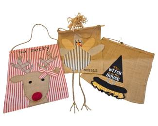 1429 - Seasonal Burlap Garden Flags Set of 3 (Description): Set of three seasonal burlap garden flags  featuring holiday-themed designs. Includes a  Christmas flag with red and white stripes and  an appliqued reindeer reading "so merry," a  Thanksgiving flag with an appliqued turkey design reading "Gobble" wiraffia hair accent,  and a Halloween flag featuring a witch's hat  design reading "The Witch is in the House"  with yellow ruffle trim. Each flag has  hanging attachments. Measures 15.5" x 8".