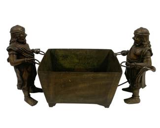 8000 - Bronze Figural Planter w/ Two Bearers (Description): Bronze figural planter featuring two  classical male figures as bearers, each  depicted in period dress with helmets and  draped garments. The figures stand on either  side of a rectangular footed vessel, holding metal rods that connect to the planter.The  piece displays a rich, dark patina consistent  with age. Measures 26 inches by 15 inches by  12 inches.