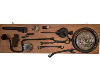 4009 - Wooden Plaque w/ Vintage Car Parts & Tools (Description): Wooden display plaque mounted with an  assortment of vintage automobile parts and  tools. The collection includes a hubcap, pipe  wrench, hand drill, various open-end  wrenches, spark plug, gauges, and other early automotive accessories and hand tools,all  arranged and mounted on a natural wood board  with hanging hardware. Measures 11" x 36".