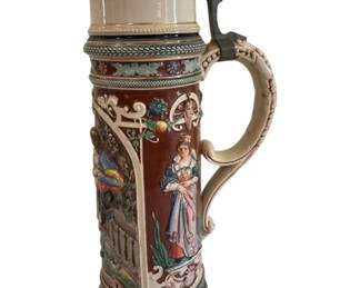 554 - German Style Ceramic Beer Stein w/ Pewter Lid (Description): Large German style ceramic beer stein  featuring a pewter lid with decorative  finial. The body displays colorful relief  scenes of figures in traditional dress set  against a rich brown background with cream-colored decorative scrollwork and  floral motifs. The ornate handle features  carved detailing, and the base is adorned  with patterned bands. Measures 16.5" tall.