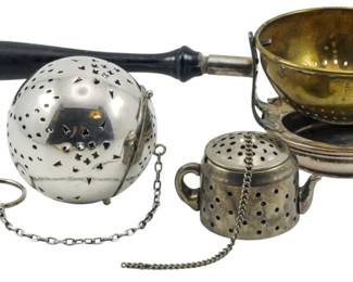 103 - 3 Vintage Tea Strainers (Description): A collection of three vintage tea strainers  featuring varied designs. The lot includes a  spherical tea ball with decorative pierced  patterns and a dark wooden handle, a small  teapot-shaped infuser with chain attachment, and a brass colander-style str