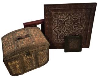 3755 - 4 Pcs Decorative Items w/ Basket & Tiles (Description): A collection of four decorative items  including a woven basket box with metal  hardware and hinged lid featuring a wooden  handle, accompanied by two large embossed  decorative wall tiles with ornate medallion designs and one smaller matching tile. The  basket displays a checkered woven pattern  with brass accents, while the tiles feature  intricate scrollwork and floral motifs in a  dark bronze-toned finish.