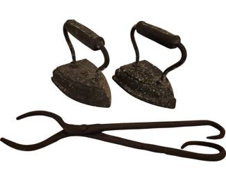 3990 - Pr Ant. Cast Iron Sad Irons w/ Hearth Tongs (Description): A pair of antique cast iron sad irons with  curved metal handles featuring wooden grips,  displaying a well-worn patina consistent with  age and use. Accompanied by a hand-forged  iron hearth tongs or ember grabber with scissor-action design and curved hooks at the  ends. These pieces are representative of  19th century domestic implements used for  pressing clothes and tending fires.