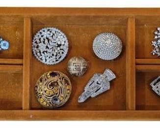 1974 - Tray w/ Paste & Rhinestone Brooches (Description): Display tray containing a collection of paste  and rhinestone brooches. The assortment  includes various styles featuring clear,  blue, and colored stones in silver and  gold-toned settings. Designs range from Art Deco geometric shapes to floral and starburst  motifs.