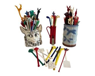 567 - Collection of Vintage Swizzle Sticks (Description): A large collection of vintage swizzle sticks  in various styles, colors, and materials. The  assortment includes figural designs  featuring animals, patriotic stars, and  novelty shapes in plastic and other materials. The lot is displayed in two  decorative holders: a whimsical cat face  ceramic container and a blue and white  bamboo-pattern ceramic vessel, along with a  miniature golf bag holder. Swizzle sticks