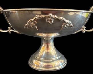546 - Sheffield Trophy Bowl 14"x10"x7" (Description): Sheffield trophy bowl featuring an elegant  pedestal base and twin scrolled handles. The  bowl displays ornate applied rococo-style  decorative cartouche on the front with  acanthus leaf and shell motifs. The piece has a polished silver-plated finish throughout.  Measures 14 inches wide by 10 inches deep by  7 inches tall.
