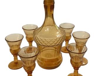 2764 - New Martinsville Amber 7pc Decanter Set (Description): New Martinsville amber glass seven-piece  decanter set. The set includes one decanter  with stopper and six coordinating stemmed  cordial glasses in matching amber coloration.