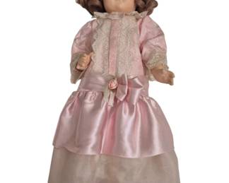1266 - Vtg Composition Doll in Pink Satin Dress 25" (Description): Vintage composition doll with brown curly  hair and painted features including red lips  and defined eyebrows. The doll is dressed in  an elegant pink satin and lace dress with  ribbon bow detail and pink rosette at the waist. She wears a matching lacehead piece  and white shoes. Displayed on a metal stand.  Measures 20".