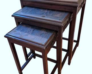 604 - Chinese Carved Rosewood Nesting Tables Set (Description): Set of three Chinese carved rosewood nesting  tables featuring intricately carved tops  depicting traditional village scenes with  figures and architecture. Each table top is  set within a raised molded frame, and the aprons display carved geometric fretwork  patterns. The tables rest on straight square  legs connected by stretchers, in classic  Ming-influenced style. The dark rosewood  finish shows beautiful patina consistent with