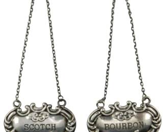 701 - Set of 2 Sterling Silver Bottle Hangers (Description): Set of two sterling silver bottle hangers  featuring ornate decorative borders and chain  attachments. One is marked 'Scotch' and the  other 'Bourbon.' Combined weight is 0.60 oz.