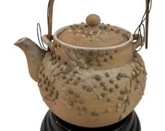 291 - Japanese Ceramic Teapot w/ Wood Base 5"x5" (Description): Japanese ceramic teapot featuring a textured  surface with raised dot and branch motifs in  a cream-colored glaze. The teapot has a woven  rattan handle and a round ball finial on the  lid. Displayed on a black lacquered wood base with stepped ring design. Measures 5  inches by 5 inches.