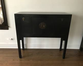 Elegant black wooden cabinet featuring clean lines and subtle Asian‑inspired design. The piece includes two upper drawers with teardrop metal pulls and two lower doors accented by a circular brass latch. Raised on four straight legs, it offers a balanced, minimalist silhouette that complements both traditional and modern interiors.  Perfect for use as a storage chest, entryway accent, or decorative display piece.  Condition: Excellent; sturdy and well maintained with minor wear consistent with age.