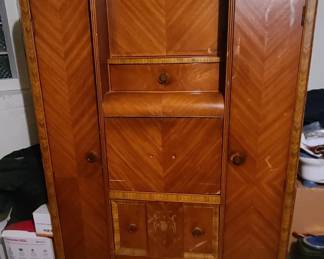 Vintage Art Deco Armoire / Wardrobe Stunning Art Deco–style armoire featuring beautiful chevron‑patterned wood veneer, rounded edges, and elegant inlay details. This substantial piece offers excellent storage with two tall side compartments and a central section that includes multiple drawers and cabinet doors. The upper corners feature curved mirrored panels, adding both charm and visual interest.  A true statement piece with classic craftsmanship and timeless style — perfect for a bedroom, dressing room, or anyone who appreciates vintage furniture with character.  Condition: Very good; well cared for with normal light wear consistent with age.