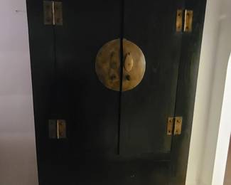 Tall Asian‑Style Wooden Cabinet with Brass Medallion Elegant dark wooden cabinet featuring a refined Asian‑inspired design with clean lines and a prominent circular brass medallion centered on the double doors. The piece includes four decorative metal hinges and a carved lower panel that adds subtle texture and craftsmanship. Its rich finish and balanced proportions make it a striking accent for any room.  Perfect for use as a storage armoire, entryway statement piece, or display cabinet.  Condition: