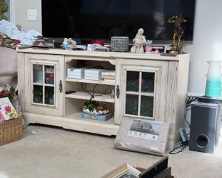 Item 4 β Distressed Cream/Antique White TV Console / Media Cabinet A charming farmhouse-style media console with a heavily distressed finish, featuring: Finish: Antique white/cream with intentional distressing revealing warm wood tones underneath β gives a well-worn, shabby chic look Doors: Two glass-panel doors (4-pane window style) with black iron-style hardware/latches, flanking a center open section Storage: Center open section with 2β3 shelves for media components; side cabinets with interior shelving Details: Fluted/reeded side pilasters, scalloped apron at base, bracket feet Style: Farmhouse / Shabby Chic / French Country Material: Wood with distressed painted finish
