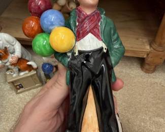 Royal Doulton "The Balloon Boy" Porcelain Figurine This appears to be the iconic Royal Doulton "The Balloon Boy" (HN 2934), one of their most beloved and recognizable figurines, featuring: Subject: A young male street vendor standing casually with hands in pockets, holding a colorful cluster of balloons Balloons: Multicolored β red, blue, yellow, green, orange, and pink β beautifully rendered in high-gloss glaze Costume: Green coat, red plaid/crosshatch neckerchief, white shirt, black trousers, brown shoes, and a cream/tan flat cap Base: Gray cobblestone-style oval base Material: Fine bone china / porcelain with hand-painted decoration Finish: High-gloss glaze throughout Condition: Appears excellent β no visible chips, cracks, or paint loss Size: Approximately 7β8" tall (standard size)
